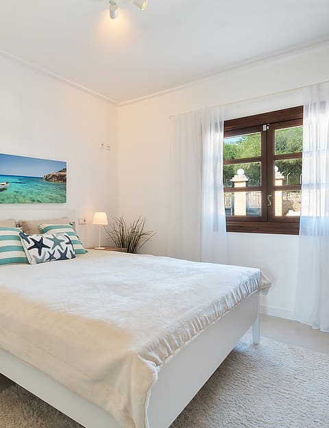 Dormitorio luminoso con cama blanca, cajoneras de madera y contraventanas oscuras con cortinas ligeras.
