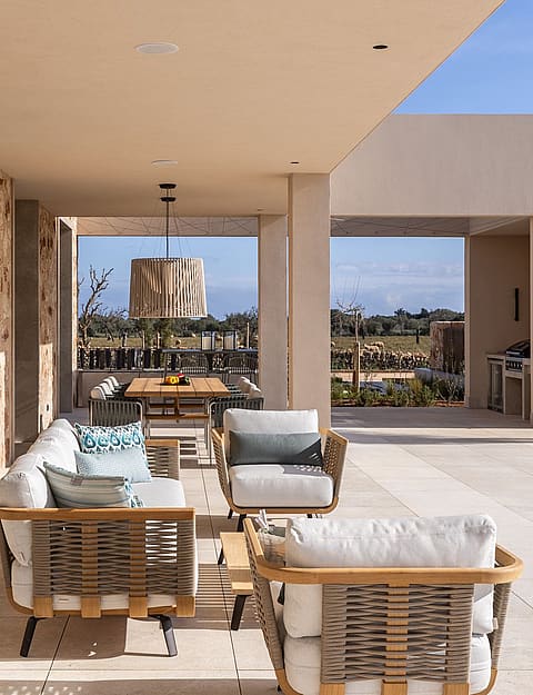 A spacious covered terrace with stone walls, outdoor lounge seating, and a dining area, overlooking a distant mountain landscape.