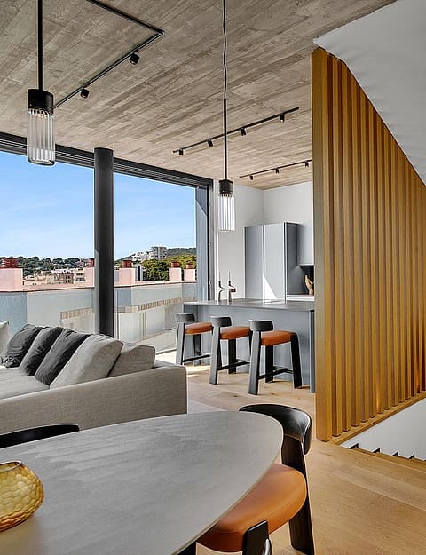 Interior of a modern penthouse featuring a wooden staircase, concrete ceiling, and open-plan kitchen with a breakfast bar.