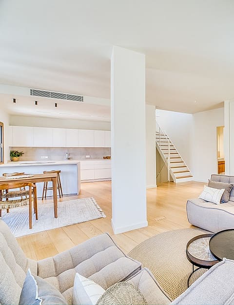 Modern open-plan living room with light oak flooring, grey tufted sofas, and a white architectural staircase.