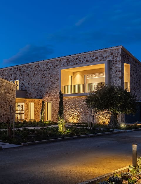 Exterior view of a modern stone-clad finca in Santanyí at twilight, showing illuminated interior spaces and landscaped grounds.