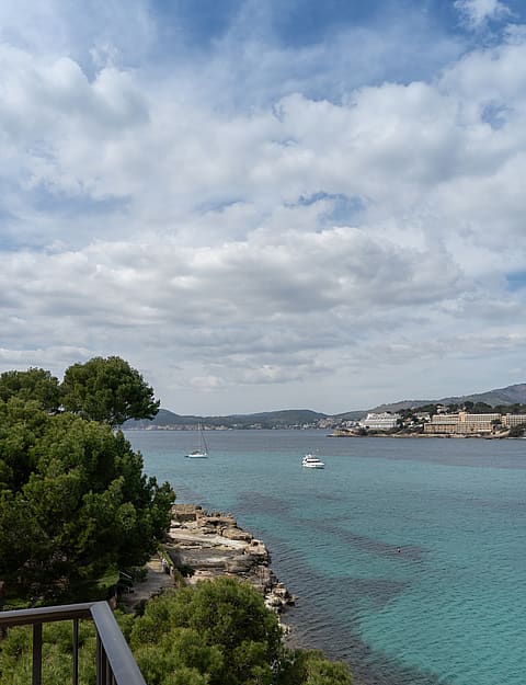 View from a terrace railing over turquoise water, a rocky shoreline, and yachts anchored in a Mediterranean bay.