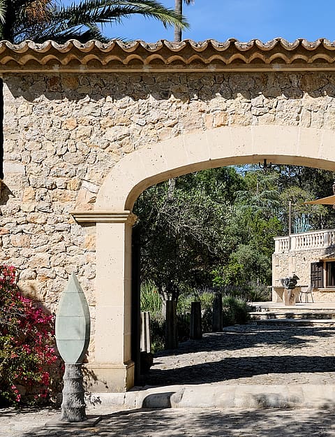 A traditional stone archway entrance to a finca, featuring rough-hewn masonry and clay roof tiles, opening to a gravel path.