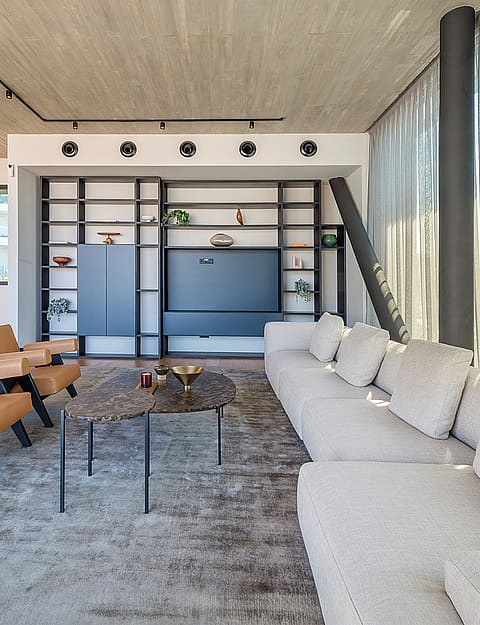 A modern open-plan living room with a textured concrete ceiling, tan leather armchairs, a large grey sofa, and a black metal shelving unit against a white wall. A wooden staircase with glass railings is visible to the left.