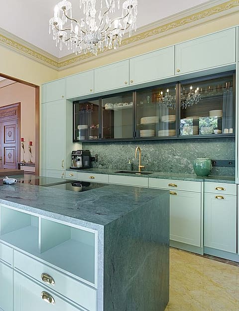 Modern kitchen with sage green cabinets, a dark grey stone island, and bar stools, leading to a dining area with a chandelier.
