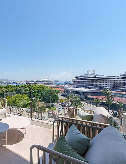 Terraza amplia con sofás grises de exterior y cojines verdes, suelo de piedra y vistas al puerto y al mar.