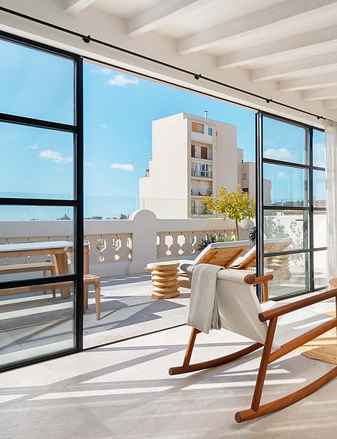 Upper-level sitting room with wooden rocking chairs and a cream sofa, steel-framed doors open to the terrace and rooftops beyond.