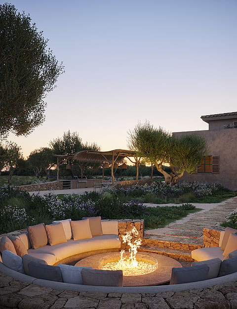 A circular stone-built fire pit with built-in seating nestled in a Mediterranean garden at dusk.