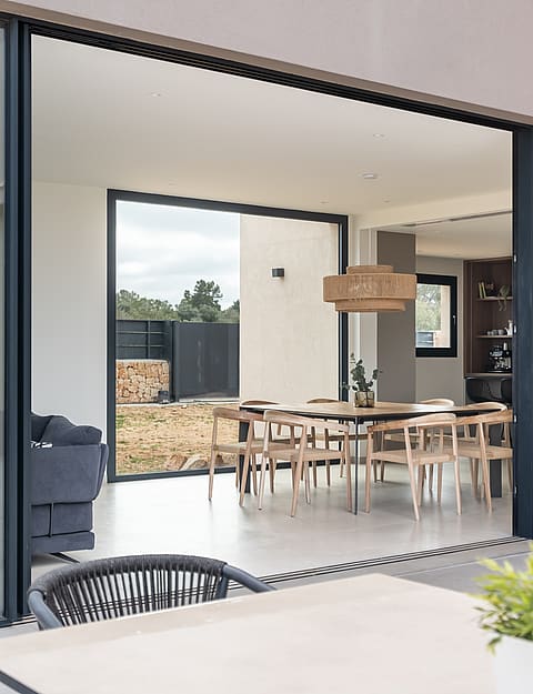 View from a terrace into an open-plan dining room with a wooden table, chairs, and large sliding glass doors.