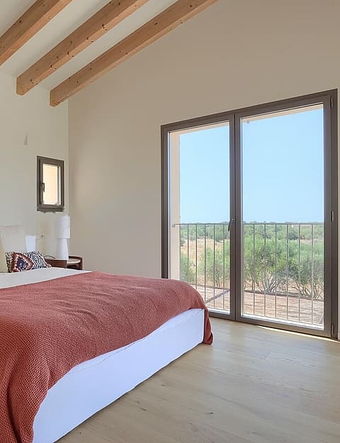 A bedroom with light wood flooring, white walls, and exposed ceiling beams. A large bed with a red cover faces a terrace door.