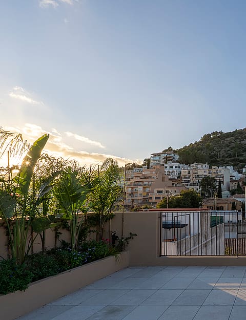 Stone-paved rooftop terrace with iron railings and planters, facing a hillside village and forested mountain peaks.