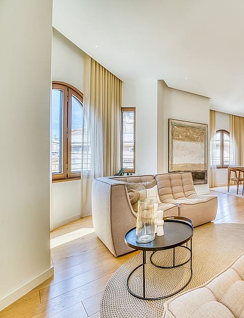 A bright living room in a penthouse with arched windows, light oak flooring, beige modular sofas, and a dining area in the background.