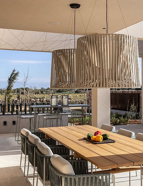 A spacious outdoor dining patio features a long wooden table, woven chairs, and a stone-built summer kitchen, looking towards mountain fields.