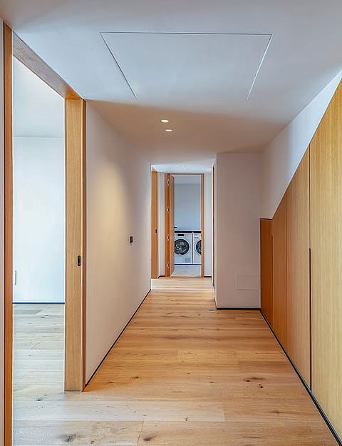 A modern residential hallway with light oak wood floors and integrated floor-to-ceiling oak wood cabinetry on the right wall.