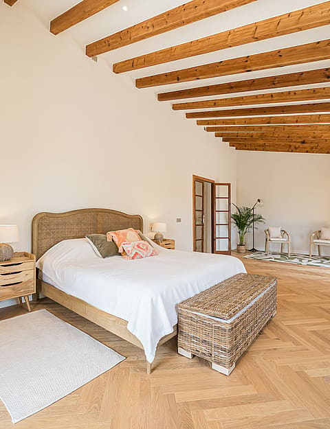 A spacious master bedroom featuring light oak herringbone floors, exposed ceiling beams, and woven furniture accents.