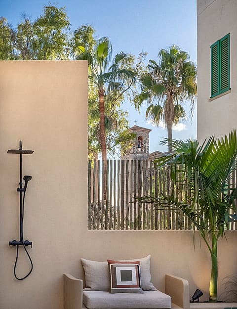 Minimalist outdoor shower with black fixtures next to a built-in bench, framed by palm trees and a village tower view.