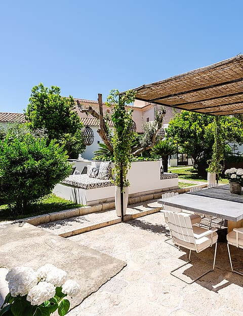 A sunny outdoor stone terrace featuring a dining table under a woven reed pergola, surrounded by lush green garden plants.