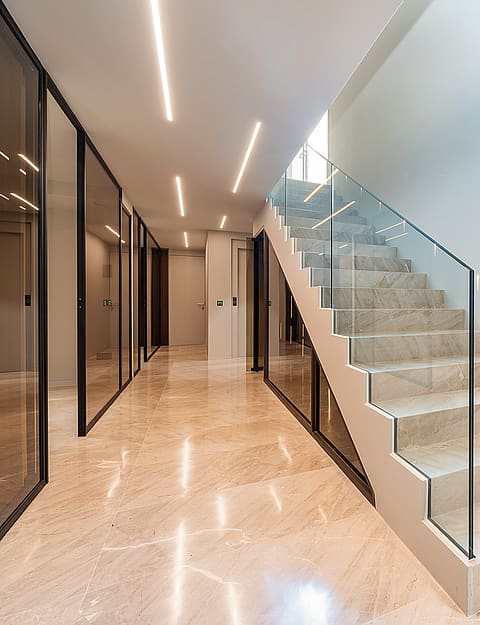 A wide interior hallway with a beige marble staircase featuring a frameless glass railing and recessed LED ceiling lighting.