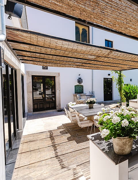 An outdoor kitchen with stone counters and a sink sits under a woven reed canopy, adjoining a paved terrace and garden.
