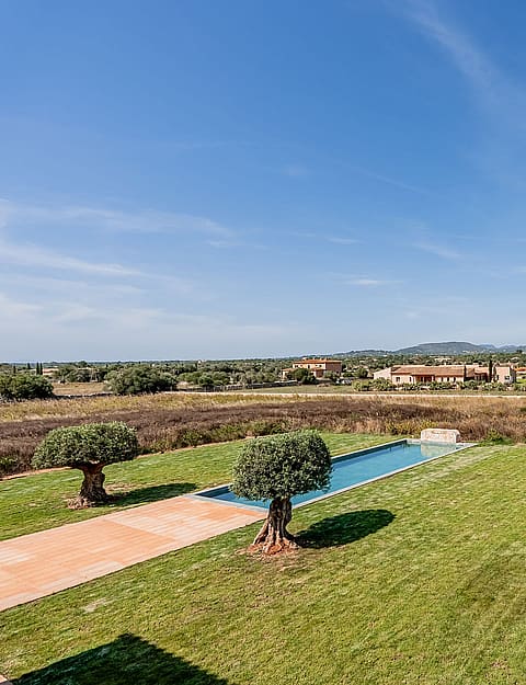 An elevated view over a green lawn and rectangular pool toward distant mountains in the Santanyí countryside.