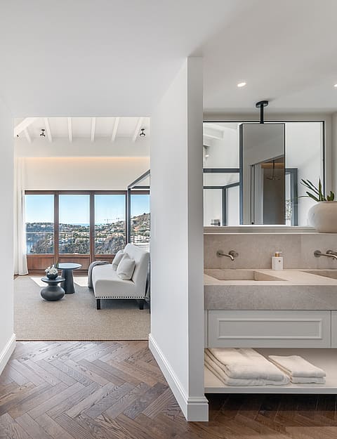 En-suite bathroom with double stone sinks and herringbone floors leading into a bedroom with a sofa and sea views.