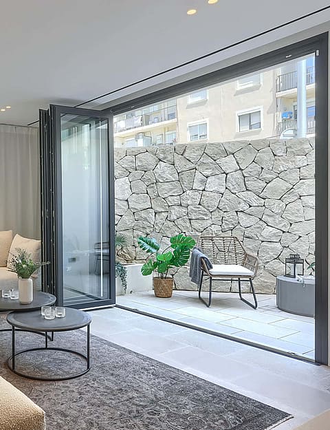 A contemporary living room with a large cream sectional sofa, dark round coffee tables, and floor-to-ceiling glass doors opening to a stone-walled courtyard.