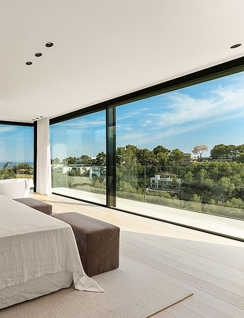 A modern master bedroom with floor-to-ceiling glass walls overlooking a wooded landscape and a distant horizon line.