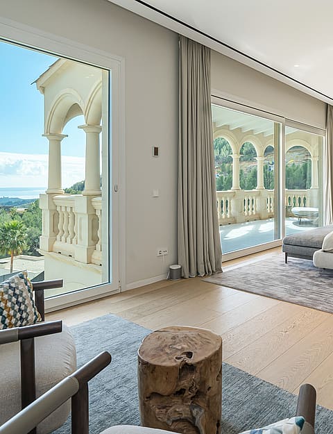 A spacious bedroom with a king-sized bed, soft grey carpet, floor-to-ceiling glass doors opening to a stone-railed balcony.