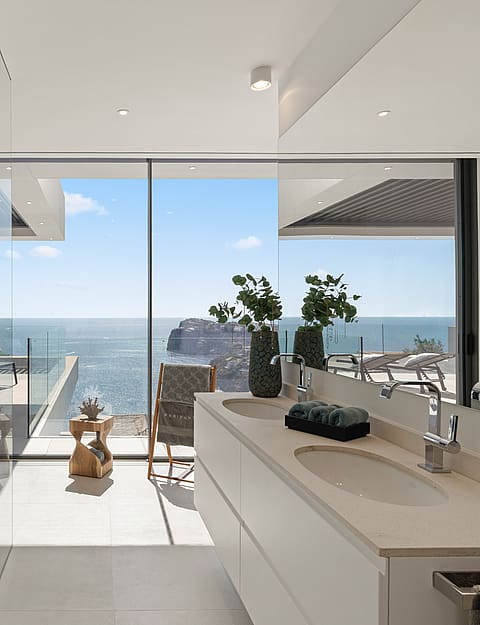 Modern bathroom featuring a double vanity, glass-enclosed shower, and floor-to-ceiling windows overlooking the ocean.