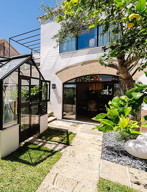 A glass and black metal greenhouse located in a private landscaped garden with a stone path and lemon tree.