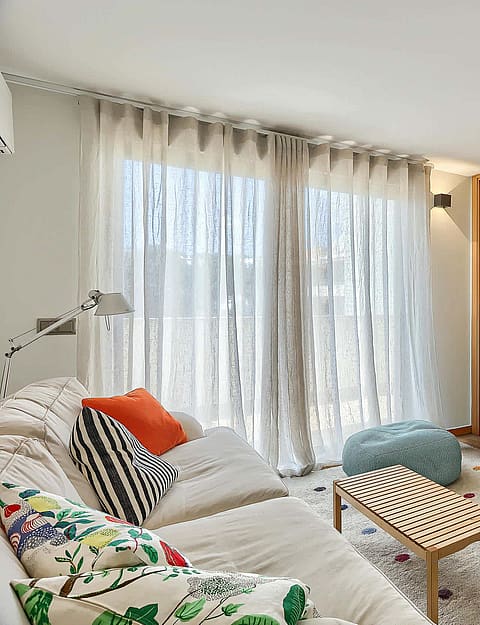 A bedroom with a double bed, large wooden floor-to-ceiling wardrobe, sheer white curtains, and a patterned area rug.
