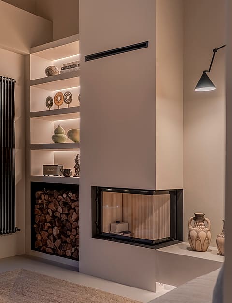 Modern glass fireplace with recessed LED shelving, stacked wood storage, and a minimalist black wall lamp.
