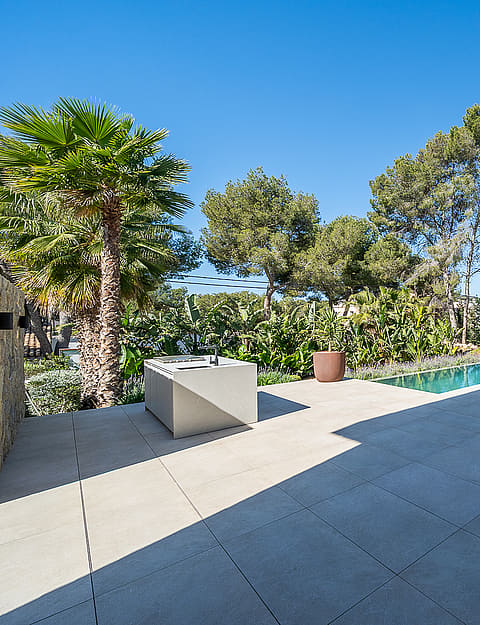 Wide view of a stone-paved terrace with a rectangular swimming pool, palm trees, and native greenery against a blue sky.