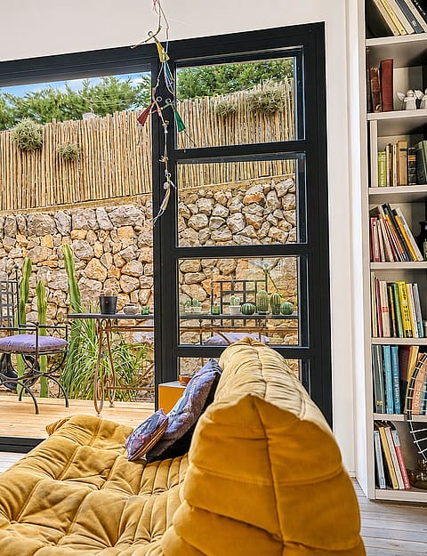 Interior living room with yellow modular sofa, glass coffee table, and large black-framed doors opening to a stone garden.