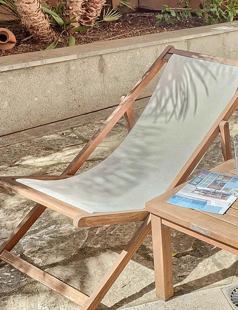 Two wooden lounge chairs with grey fabric seats arranged on a stone patio beside a small square side table with drinks.