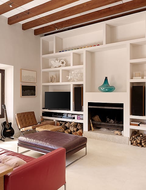 Living room with exposed wood ceiling beams, white built-in shelving unit, dark red sofa, and an open fireplace.