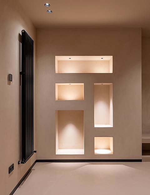 A smooth beige interior wall with four asymmetrical recessed alcoves, each illuminated by warm integrated spotlights, next to a black vertical radiator.