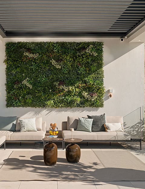 A shaded outdoor lounge area featuring a large vertical garden wall, cream-colored modular sofas, and stone side tables.