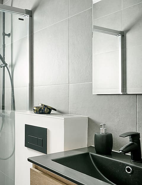 A bathroom with grey tiled walls, a glass-enclosed shower with a rainfall head, and a vanity with a black sink.