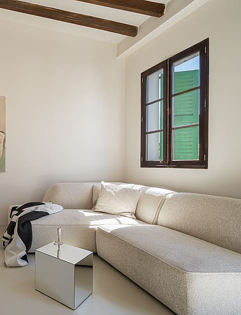 Minimalist living area featuring microcement flooring, light textured sofa, exposed timber beams and a large potted plant.