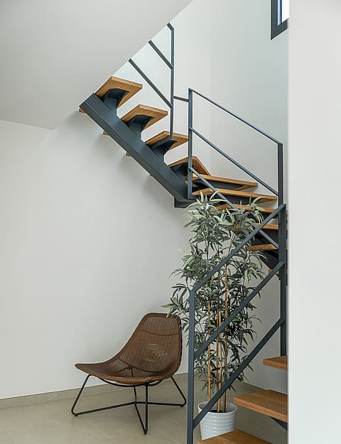 A modern staircase with dark metal frame, oak steps, and a white interior, featuring a woven accent chair underneath.