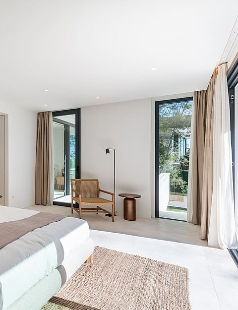 Serene master bedroom with neutral linen bedding, rattan accent chair and floor-to-ceiling sliding doors to a private terrace with pine tree views