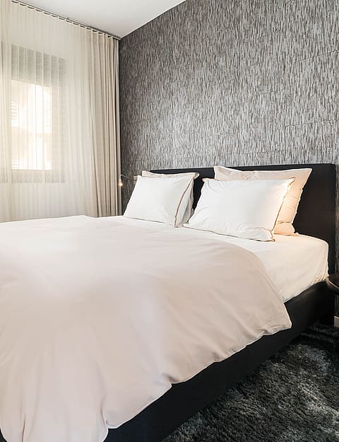 A modern bedroom with a black upholstered bed frame, light bedding, and a grey vertical-patterned accent wall. Sheer curtains cover the window to the left.