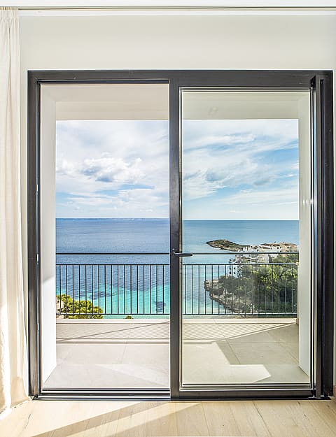 A sliding glass door framed by cream curtains leads from a bedroom to a sunlit terrace overlooking the Mediterranean Sea.
