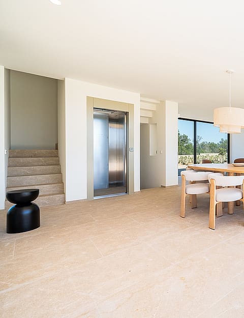Interior view of an open-plan kitchen and dining area featuring a light wood dining table, cream chairs, a central home elevator, and floor-to-ceiling glass doors.
