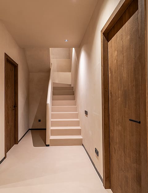 A neutral-toned hallway in a Genova townhouse featuring a wooden shelving unit with wicker baskets, solid wood doors, and a limestone staircase leading upwards under soft recessed lighting.