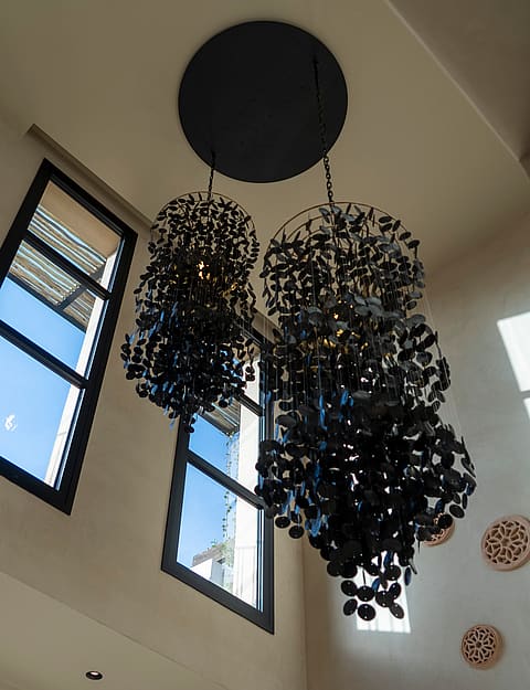 A low-angle shot of two dark, intricate, floral-inspired pendant light fixtures hanging from a high ceiling near tall windows.
