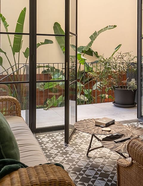 A living space with rattan furniture and patterned floor tiles opening through black-framed glass doors onto a green terrace.