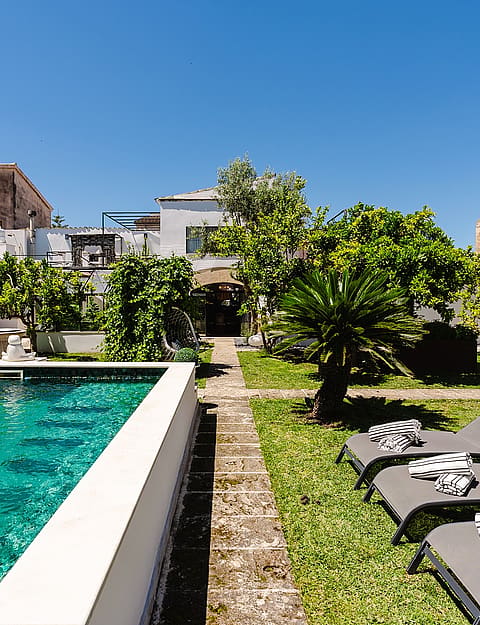 A rectangular swimming pool with turquoise water bordered by a stone path and a grassy lawn with three sun loungers.
