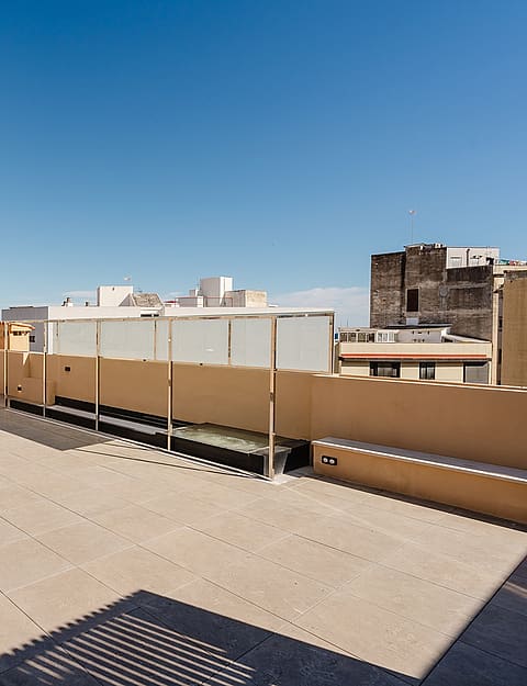 A wide, empty rooftop terrace paved with light tiles, featuring low perimeter walls, glass balustrades, and distant city buildings under a clear blue sky.